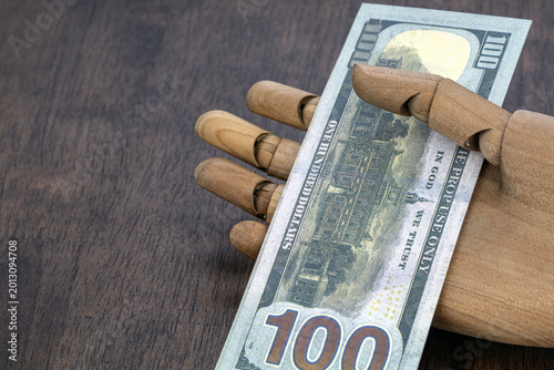 Wooden articulated artist mannequin hand holding a US one hundred dollar bill, showing the back side with Independence Hall and In God We Trust inscription on dark wooden surface