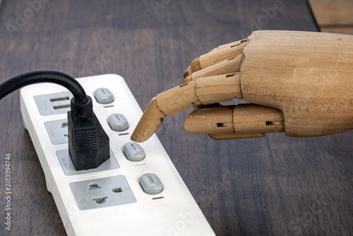 Close-up of a jointed wooden robotic hand with extended index finger pressing a gray button on a white multi-outlet power strip surge protector placed on dark wooden surface