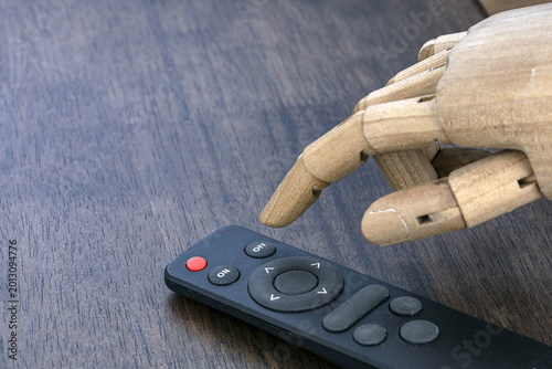 Articulated wooden mannequin hand reaching to press button on black TV remote control placed on dark wooden table surface, concept of AI automation and technology