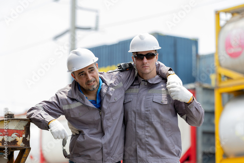 Fotografía Two construction workers wearing helmets and gloves, supporting each other with arms around shoulders at an industrial site