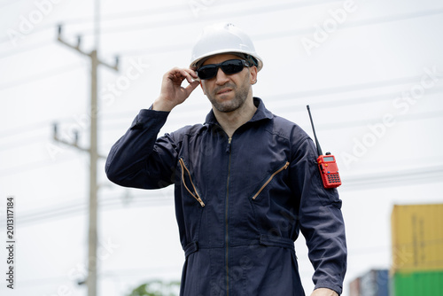 Foto A man in a safety helmet and dark coveralls adjusts his sunglasses outdoors near power lines, wearing a red walkie-talkie on his arm