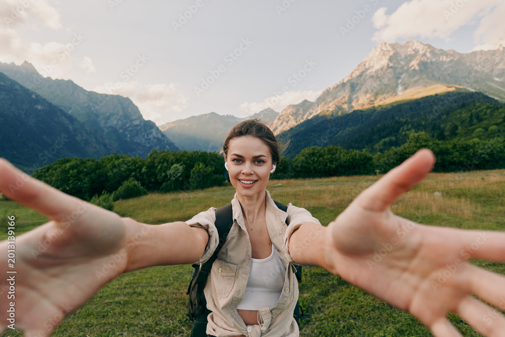 Obraz premium Cheerful woman outdoors, smiling toward the camera, arms extended, sunlit meadow with trees, distant mountains, casual outfit, natural colors, lifestyle moment