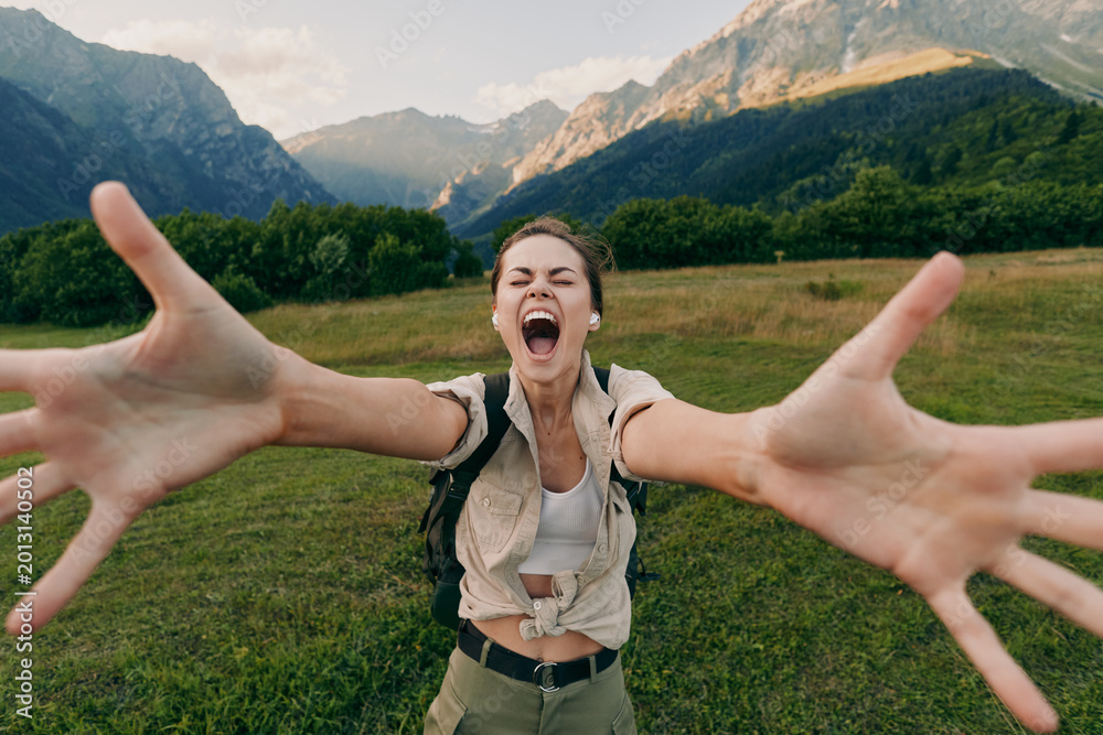 Fototapeta premium Happy woman with backpack stretching arms towards camera in green meadow with mountains in background. Outdoor lifestyle, adventure, travel, and freedom concept.