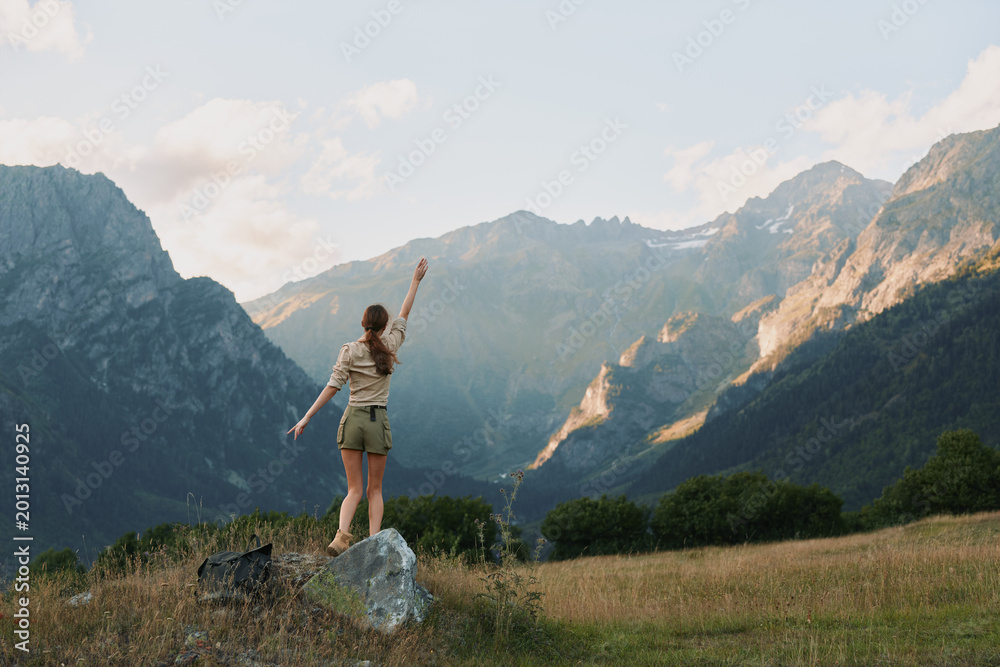 Fototapeta premium Woman outdoors on a rock in a meadow, arms raised in a carefree pose, majestic mountains in the background, sunlit scene, natural colors, lifestyle moment