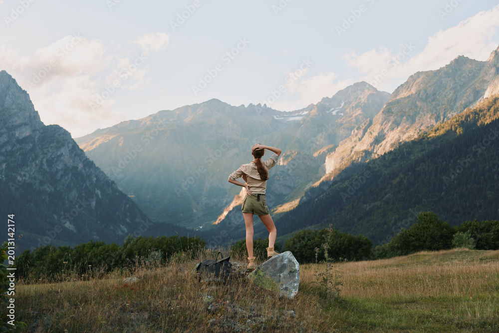 Naklejka premium Young woman stands on a rock in a sunlit valley, arms raised in triumph as distant mountain peaks glow under a clear blue sky, wearing beige tank top and light shorts, embodying freedom