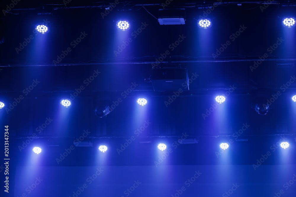 Fototapeta premium Cool blue LED stage lights shine through haze from a dark ceiling rig, creating a moody concert or theater background with copy space