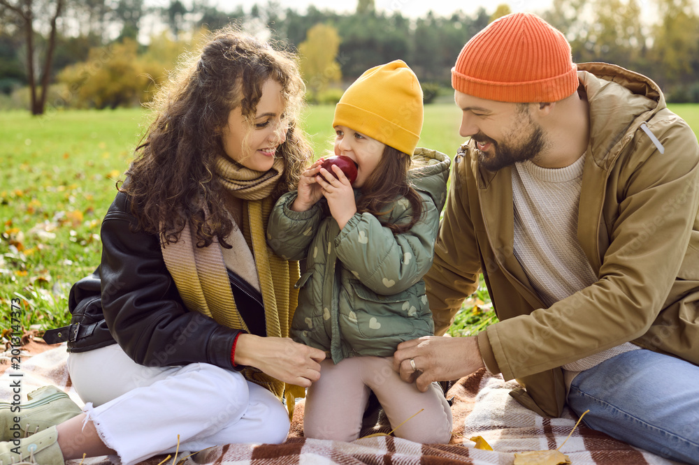 Fototapeta premium Family enjoys autumn picnic with happy daughter. Parents relax on a blanket in the park as their child enjoys a red apple, bundled in warm clothing. Happy family time outdoors in sunlight.