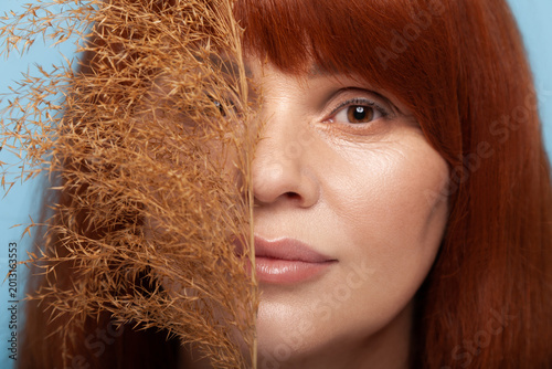 Portrait of attractive redhead woman posing with dry decorative grass branch near face for aesthetic beauty photography session isolated on blue background