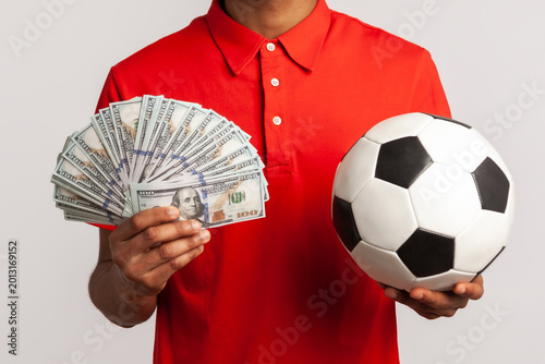 Smiling satisfied man with dreadlocks wearing red casual style T-shirt, holding soccer ball and fan of dollar bills, sports betting, big win. Indoor studio shot isolated on gray background.