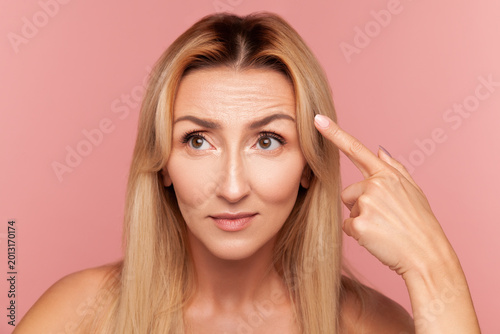 Concerned blonde lady pointing at her forehead skin texture while checking for fine lines and aging signs during beauty routine isolated on pink background