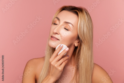 Middle aged woman massaging her chin with soft white cotton pad for deep skin cleaning and aesthetic hydration treatment isolated on pink background