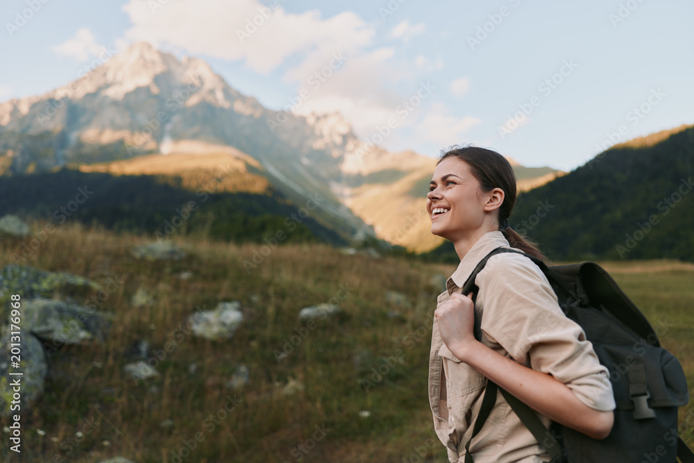 Fototapeta premium Woman hiking outdoors with a backpack, smiling as she walks through a sunlit meadow, with distant mountains and lush scenery, showcasing a relaxed lifestyle.