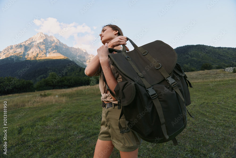 Naklejka premium A woman with a large backpack stands in a meadow with mountains in the background, preparing for a hike. An outdoorsy lifestyle moment emphasizing adventure and travel.