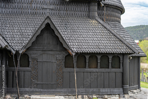 Traditional Norwegian stave church surrounded by spring countryside and forested hills under dramatic cloudy sky in Telemark, Norway. Dark wooden medieval-style architecture. 