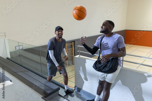 Obraz na plátne Diverse male friends tossing basketball and reaching on bleacher steps at gym wi
