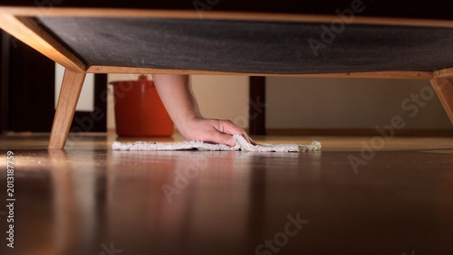 Konstfotografi Hand reaching with a rag under dark furniture to wipe dusty hardwood floor, clos