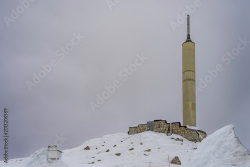 Winter view of the iconic communication tower and mountain facilities on the summit of Gaustatoppen. Snow-covered rocky terrain under a dramatic overcast sky in the Norwegian mountains. 