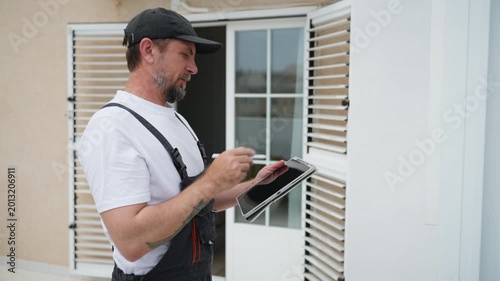 Handyman in overalls inspecting window shutters and taking notes on tablet. Male contractor preparing detailed work estimate and renovation budget for client.