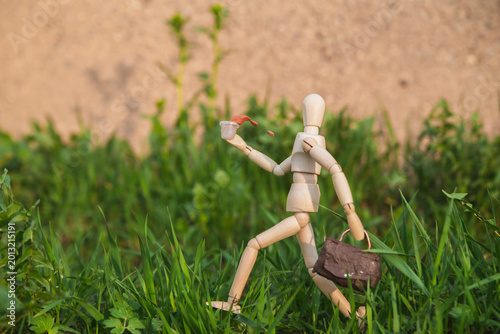 Bild auf Leinwand Wooden Figure of Man Carrying Brown Bag Walking On Grass Holding Cup Of Spilling