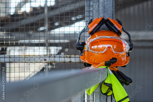 Tablou pe pânză Orange hard hat with attached earmuffs and safety goggles on a yellow high visibility safety vest hanging on metal railing at construction site