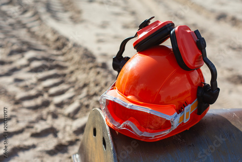 Photography Orange hard hat with attached ear muffs and protective goggles resting on a metal pipe at a sandy construction site