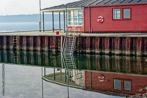 Photography Calm waterfront scene with red boathouse and metal ladder reflecting in still wa