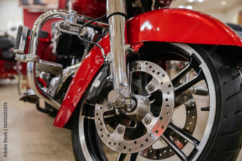 Fototapeta premium Detailed view highlights front wheel, tire, brake disc, and chrome components of red motorcycle. Low-angle perspective emphasizes robust engineering and polished surfaces in showroom setting.