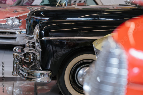 Close-up shot highlights vintage black car's chrome grille, bumper, and whitewall tire. Other classic cars, including pink and red models, are visible in the polished showroom background.