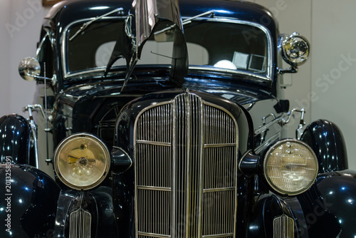 Detailed front view of classic dark blue automobile showcasing chrome grille, circular headlights, and partially open hood. Indoor setting highlights the car's glossy finish and elegant design.