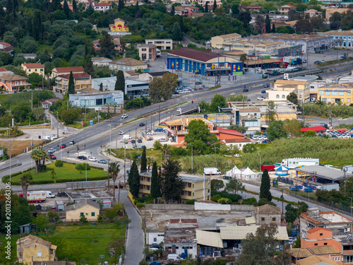 Aerial view of a busy intersection in Corfu, Greece, showing a supermarket, amusement park, local traffic, and suburban infrastructure surrounded by nature.