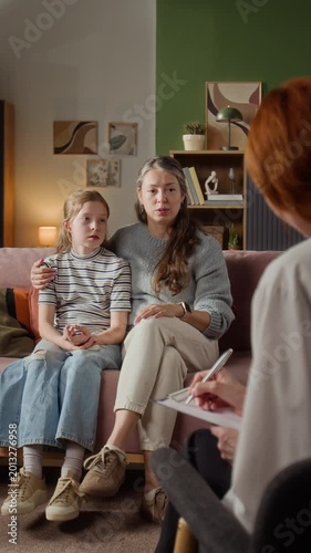 Vertical shot of adult mother discussing young daughters cough and fever symptoms with female pediatrician while doctor taking notes during house call
