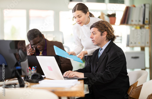 Fotografia Woman with documents oversees work of male subordinate, looks at laptop screen and helps to make proposal for counteragents