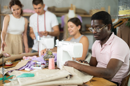 Фотография Focused middle-aged man sewing while others working with sewing-machine and maki