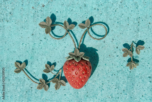 A decorative metal strawberry ornament with intricate leaf motifs set on a turquoise textured surface, creating a playful still-life composition that evokes summer, craftwork, and vintage charm.