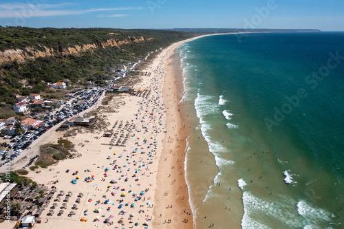 Aerial drone shot of Praia de Fonte da Telha beach in Costa de Caparica, Portugal. Sunbathers and swimmers enjoy the packed beach while the parking lot fills
