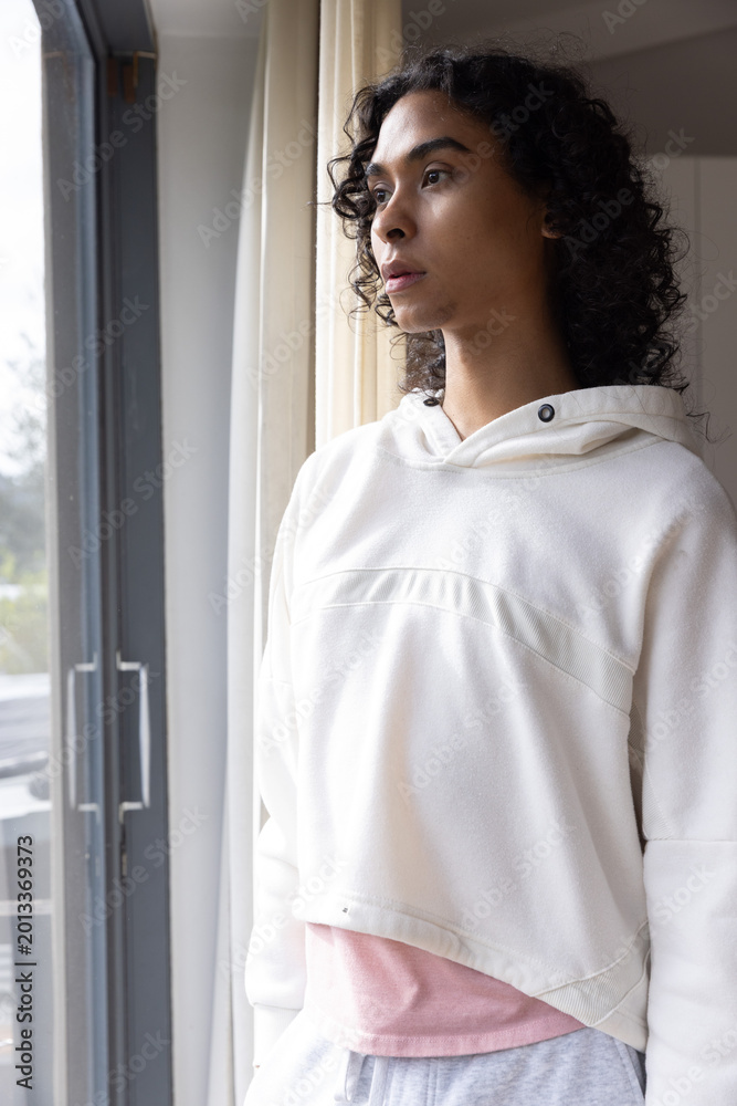 Fototapeta premium Nonbinary person wearing hoodie standing, gazing out sliding door inside with partly drawn curtain