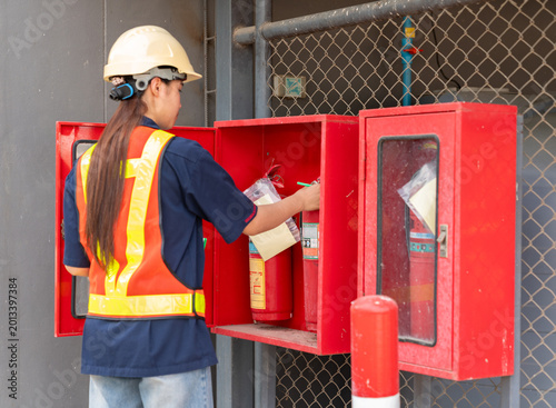Female safety officer or inspector in a hard hat and reflective vest performing a maintenance check on fire extinguishers in a red storage cabinet at an industrial site.