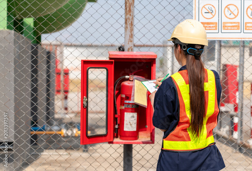 Female safety officer in a hard hat and reflective vest inspecting a fire extinguisher and recording the maintenance status on a checklist at an industrial facility.