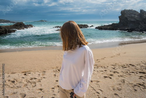 Tableau sur toile Woman from behind gaze at ocean waves, rocky beach, and dynamic sky on a peaceful day
