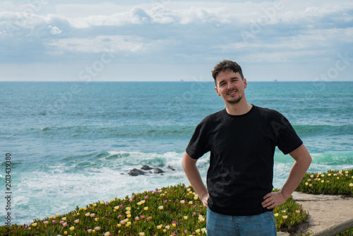 Canvastavla Confident Young Man Posing Against Dynamic Ocean Backdrop with Coastal Flowers on a Bright Day
