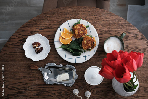 A stylish breakfast spread on a wooden table: crispy fritters with greens and orange slices, a cup of frothy coffee, dates on a decorative plate, butter dish, and a vase with vibrant red tulips.