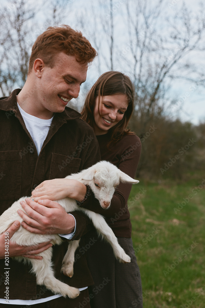 Fototapeta premium A joyful couple outdoors holds a fluffy lamb, smiling as they cradle the young sheep in a green field, capturing a candid lifestyle moment of gentle animal care and love