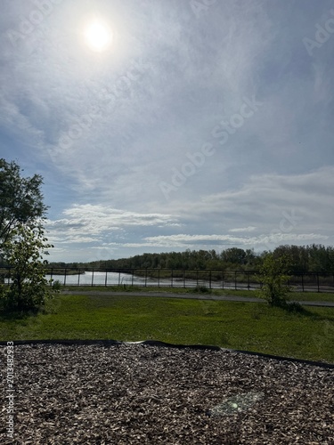 Sunlit Grassy Park With Gravel Path Overlooking Calm Lake And Wooden Fence, Scattered Trees Near Horizon, Soft Cloud Cover And Radiant Sun Creating Tranquil Atmosphere Ideal
