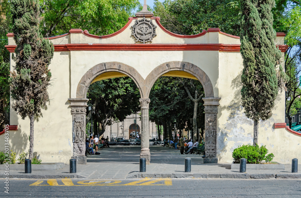 Fototapeta premium Arco de entrada colonial al atrio de San Juan Bautista en Coyoacán, Ciudad de México, con ambiente de parque