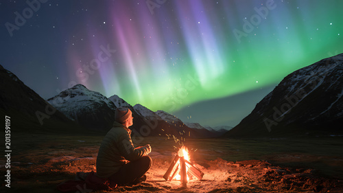 Man sitting by campfire, watching Northern Lights, aurora borealis display, mountain landscape