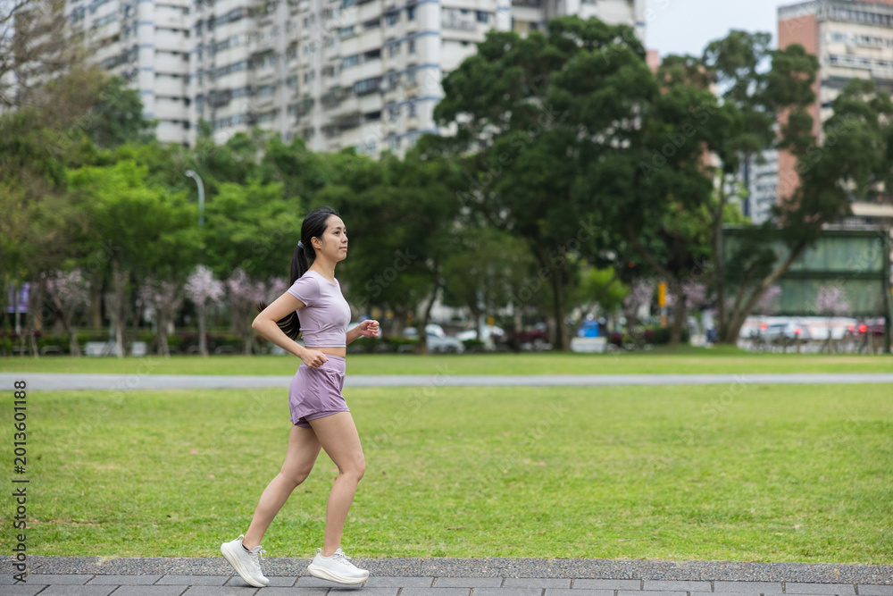 Fototapeta premium Active young asian woman jogging through a green urban park during morning workout
