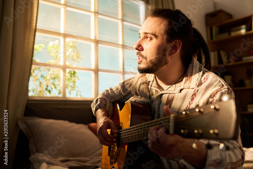 Young adult Caucasian man playing acoustic guitar and singing at home, practicing music and songwriting. Useful for creative hobby, home entertainment, musician lifestyle marketing
