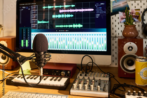 Close up of podcast microphone recording audio beside computer editing waveform tracks in home studio. Supporting content creation, voiceover production, streaming, marketing media