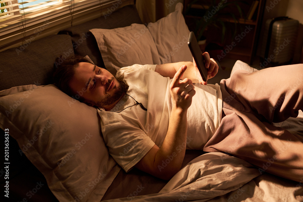 Fototapeta premium Young adult Caucasian man lying in bed, using smartphone at night, scrolling before sleep and showing screen time habit. Useful for sleep disruption, mobile addiction, bedtime routine themes