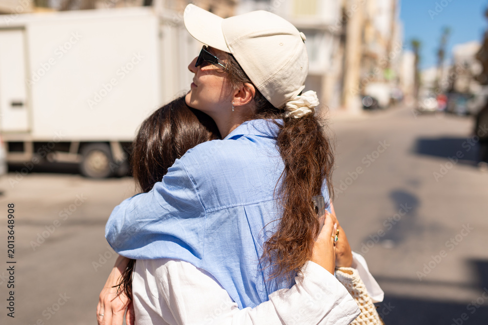 Fototapeta premium Two women are hugging each other on a sunlit city street. One woman is wearing a blue shirt and a beige cap, while the other is in a white top.